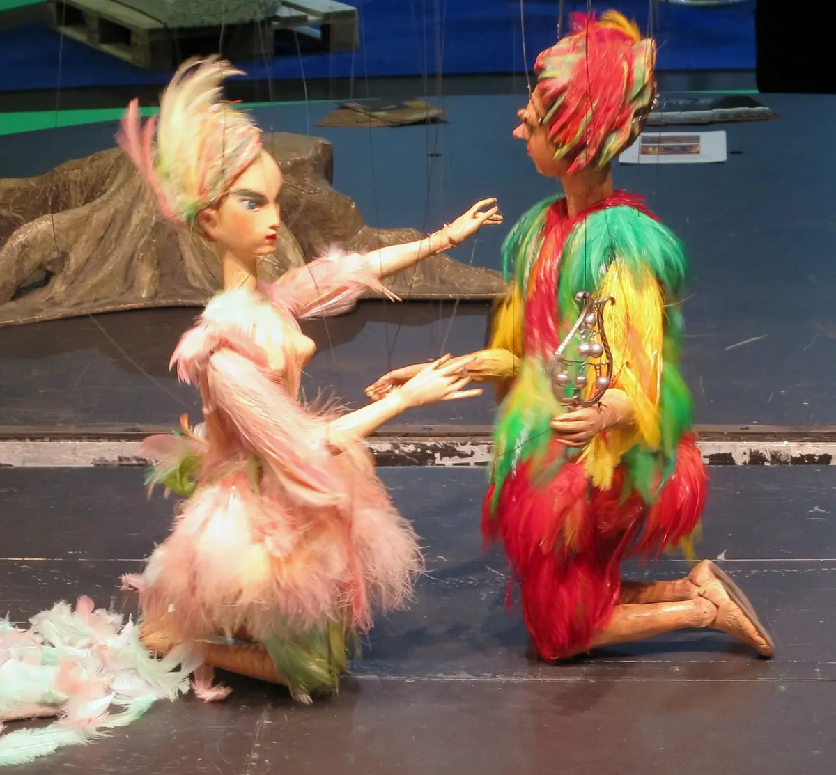 Dwie marionetki w strojach z piór, jedna różowa, druga czerwono-żółto-zielona, klęczą naprzeciwko siebie na scenie, a nad nimi widoczne są cienkie sznurki