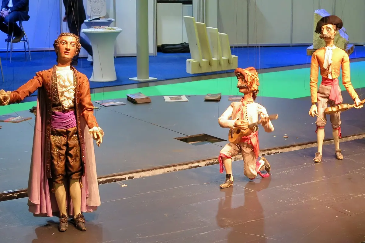 Tre marionetter i attenhundredetalskostumer står på en nøgen scene: en høj figur i kappe med lilla foer, en knælende figur med lut og en stående figur med hat